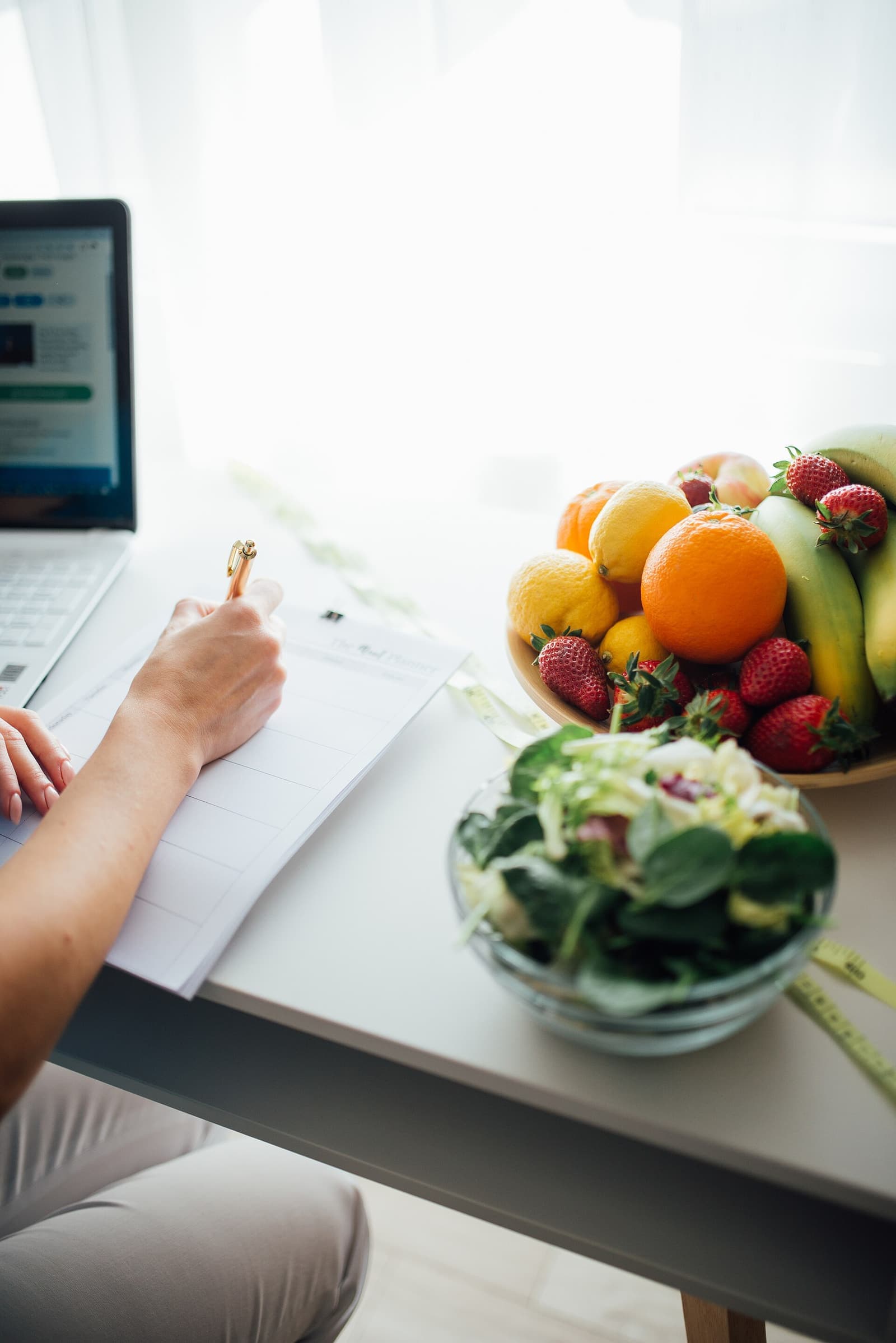 Mão anotando um plano alimentar ao lado de frutas e salada